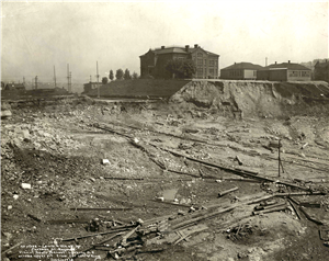 A large brick school building, and small wooden buildings, perched on a muddy bluff below which is a vast valley of mud and debris